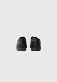 Et par svarte skinn loafers med tykke gummisåler, sett bakfra på en enkel hvit bakgrunn.