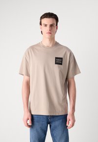 Tricou din bumbac bej cu mâneci scurte, guler rotund și un buzunar dreptunghiular negru pe piept. Textul alb de pe buzunar spune "BARBOUR ENGLAND."