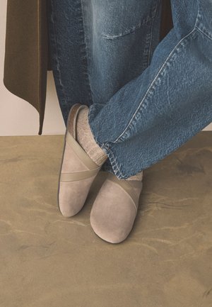 Osoba nosząca beżowe buty wsuwane, jasnobrązowe skarpetki, niebieskie dżinsy, stojąca na beżowej teksturowanej podłodze obok brązowego płaszcza.