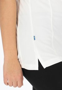 Hvid træningstop lavet af åndbart stof, med en sideslids og en blå-gul label i kanten. Kombineret med sorte leggings.