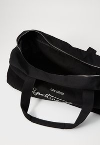 Bolsa de lona negra con cierre de cremallera, que cuenta con dos asas resistentes y una impresión en texto blanco "LES DEUX Sporting" en un lado. El interior no está forrado.