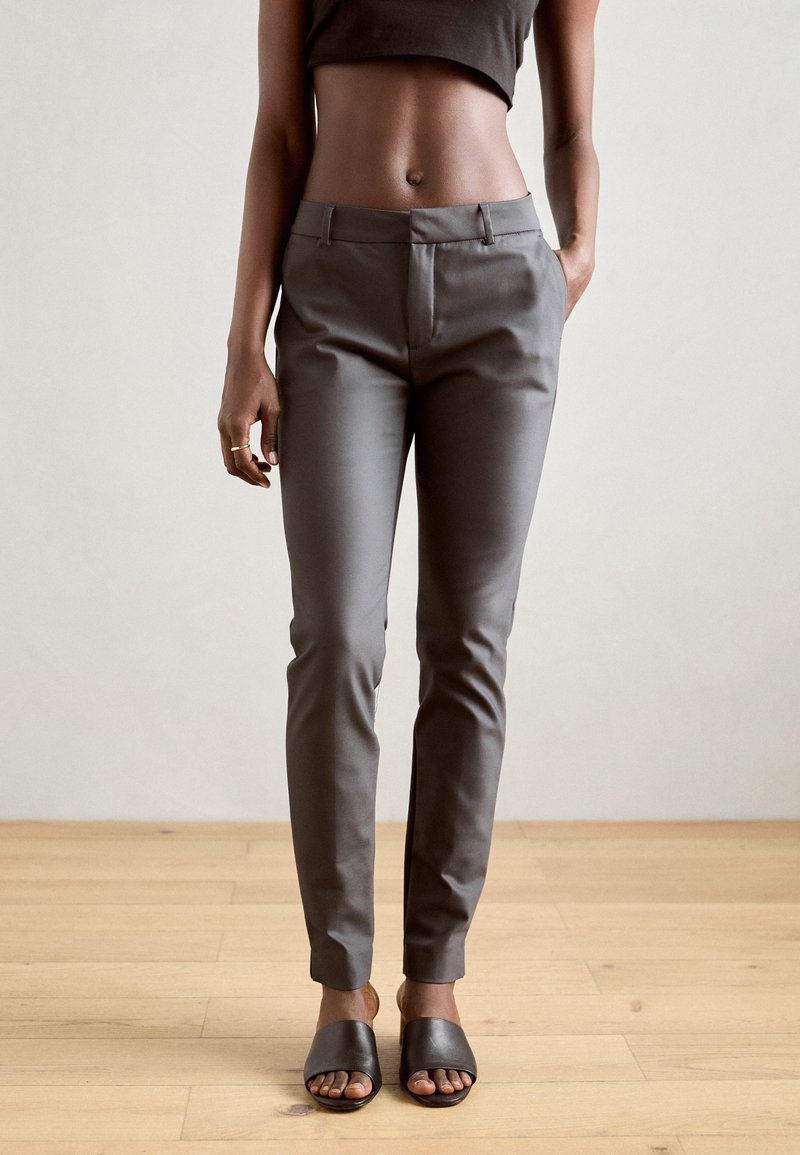 Femme portant un pantalon ajusté gris foncé, un top court noir et des sandales noires à bouts ouverts, debout sur un sol en bois.