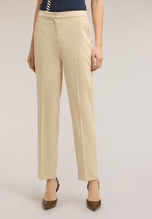 Donna che indossa pantaloni beige a gamba dritta e tacchi marroni con punta a punta, in piedi contro uno sfondo chiaro e uniforme.