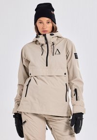 Beige ski-jack met een halve rits aan de voorkant, verstelbare capuchon, zwarte ritsen en een logo. Gecombineerd met zwarte handschoenen en een gebreide muts.