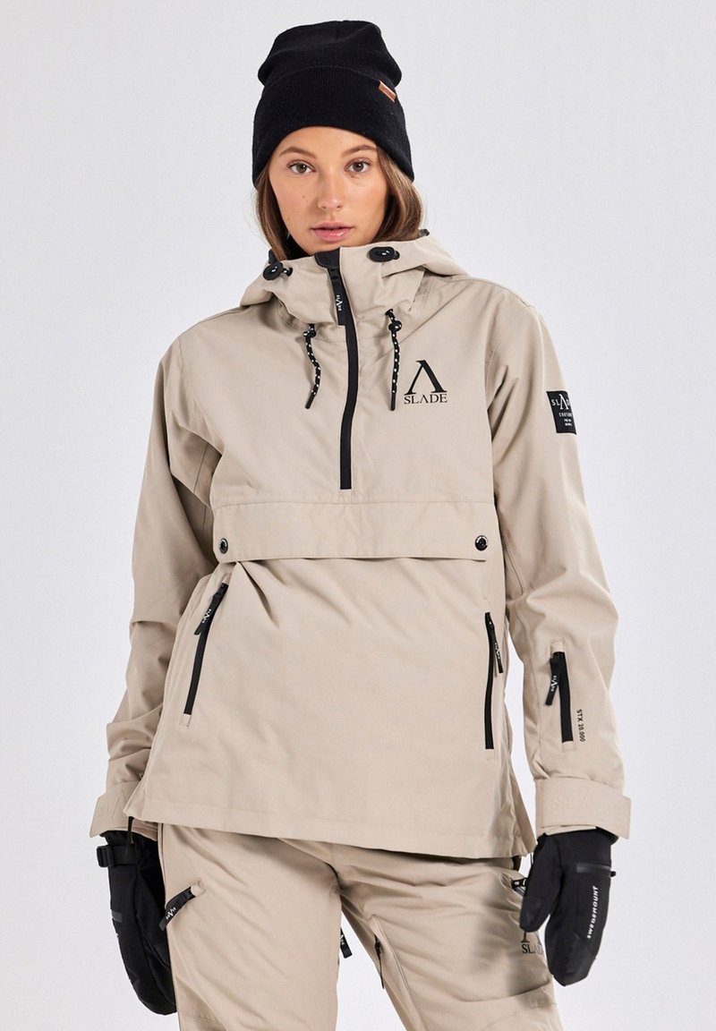 Beige ski-jack met een halve rits aan de voorkant, verstelbare capuchon, zwarte ritsen en een logo. Gecombineerd met zwarte handschoenen en een gebreide muts.