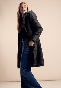 Zwarte gewatteerde jas met capuchon, met een rits aan de voorkant en een gestructureerde stof. Gecombineerd met blauwe jeans met hoge taille, die een rechte pasvorm hebben.