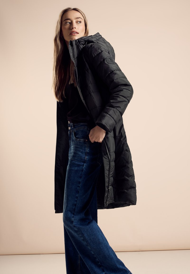 Zwarte gewatteerde jas met capuchon, met een rits aan de voorkant en een gestructureerde stof. Gecombineerd met blauwe jeans met hoge taille, die een rechte pasvorm hebben.