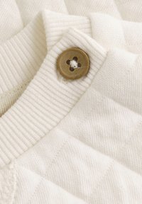 Tissu en tricot blanc avec un col côtelé et un bouton marron à quatre trous. Motif texturé visible sur le tissu.