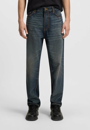 Personne portant un jean bleu foncé délavé et des baskets noires, se tenant devant un fond clair uni.