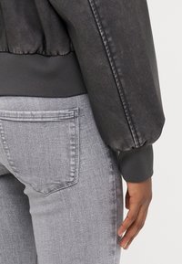 Svart bomberjakke med elastiske mansjetter, parret med lysegrå jeans med baklomme. Jevn tekstur, tettsittende design, minimale detaljer.