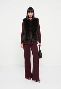 Gilet en fausse fourrure avec des bordures marron, porté sur un costume bordeaux ; des pantalons larges, des talons ouverts et un petit sac à main noir complètent le look.