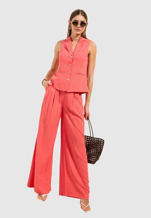 Frau trägt eine korallenfarbene ärmellose Weste mit Knöpfen und weit geschnittene Hosen, hält eine braune gewebte Handtasche, trägt Sonnenbrille und weiße Sandalen.