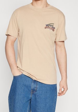 Camiseta de algodón beige de manga corta, con un gráfico colorido de "Jeans" en el lado izquierdo del pecho. Combinada con jeans de mezclilla azul.