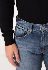 Jeans din denim albastru au o talie medie, cu nituri din cupru, buzunare cusute și un aspect decolorat. Sunt asortați cu un top negru cu mânecă lungă.
