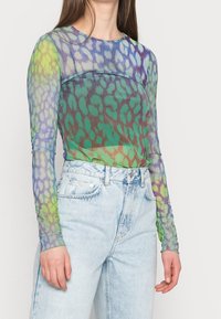 Durchsichtige Langarmbluse in Grün, Blau und Lila mit einem Leopardenmuster. Kombiniert mit hellblauen, hochgeschnittenen Jeans.