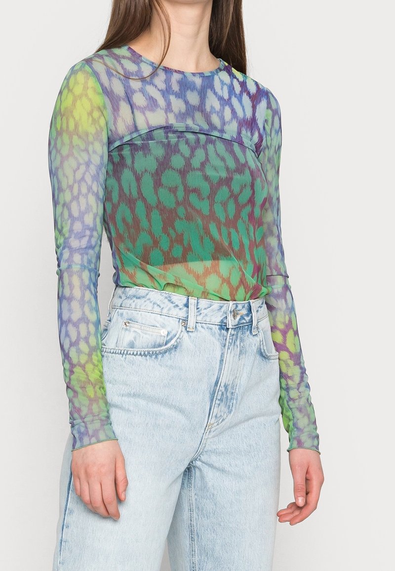 Durchsichtige Langarmbluse in Grün, Blau und Lila mit einem Leopardenmuster. Kombiniert mit hellblauen, hochgeschnittenen Jeans.