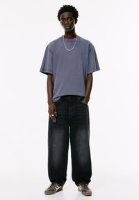 Ung mand iført overdimensionerede, falmede sorte jeans, løs grå t-shirt, lagdelte sølv kæder, ringe og sneakers, står med hænderne i lommerne.