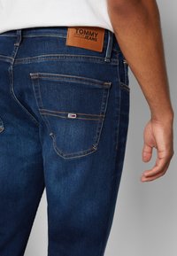 Modré denimové džíny se záběrem na zadní kapsu, která obsahuje hnědou koženou visačku s nápisem "TOMMY JEANS" a kontrastní oranžové švy.