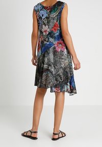 Monivärinen hihaton mekko, jossa kukka- ja eläinkuviot, laskeutuva kangas, polvipituinen ja epäsymmetrinen helma, yhdistetty mustiin sandaaleihin.