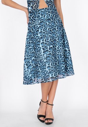 Kobieta ubrana w niebiesko-czarną midi spódnicę w panterkę, zestawioną z czarnymi sandałami na wysokim obcasie z paskiem wokół kostki, stojąca na białym tle.