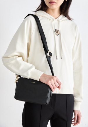 Persona con sudadera con capucha crema y pantalones negros sosteniendo un pequeño bolso bandolera negro con una correa ajustable en el hombro.
