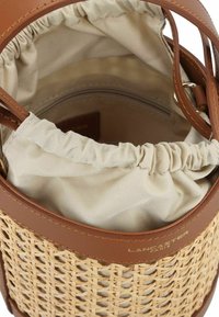 Sac seau en paille tissée avec des finitions en cuir brun. Il comprend une doublure en tissu beige froncée et une poche zippée à l'intérieur.