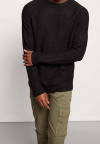 Homme portant un pull noir en maille côtelée et un pantalon cargo vert olive, debout avec un bras croisé sur l'autre.