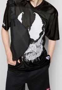 Black mesh jersey with white graphic featuring a skull design. Black crossbody bag with zippers, worn over the shoulder, paired with dark pants.