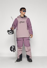 Paars en grijs snowboardoutfit met een capuchon, ritszakken en zwarte accenten. Model houdt een snowboard vast en draagt een zonnebril.