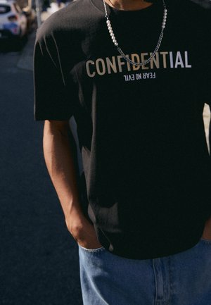 Person wearing a black t-shirt with "CONFIDENTIAL" and upside-down text "FEAR NO EVIL," a mixed pearl and chain necklace, with hands in blue jeans pockets.