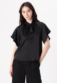 Blusa nera a maniche corte con scollo a fiocco, texture morbida, bordi ondulati e una vestibilità rilassata, abbinata a pantaloni neri.