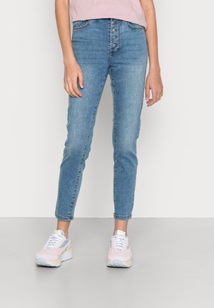 Jeans de cintura alta azul claro com botão na frente, com um corte slim, textura desbotada e comprimento até ao tornozelo. Usados com ténis em tons pastel.