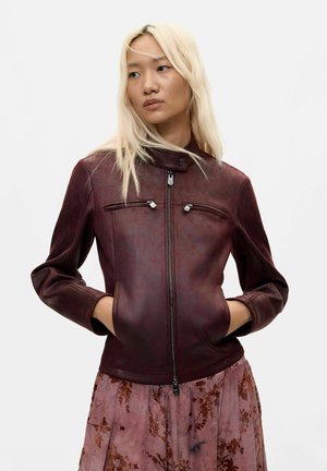 Femme aux longs cheveux blonds portant une veste en cuir rouge foncé et une jupe fleurie rose, les mains dans les poches de la veste, regardant vers sa gauche.