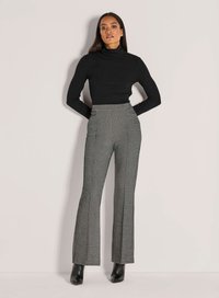 Vrouw in een zwarte geribde coltrui, hoge taille zwart-wit gestreepte broek en zwarte enkellaarzen, staand tegen een lichte muur.