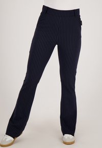 Navyblauwe pinstripe broeken met een slim fit en iets uitlopende pijpen, gedragen met witte sneakers op een witte achtergrond.