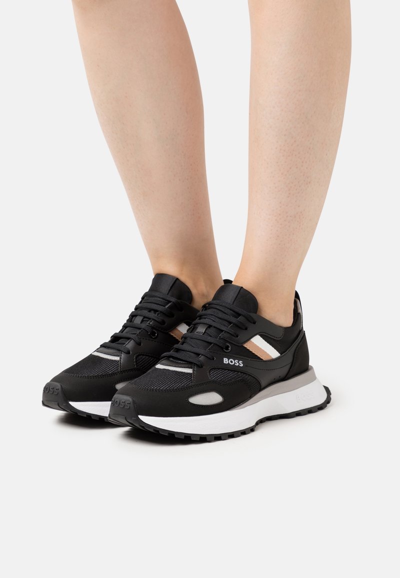 Baskets de sport noires avec tige en mesh et synthétique, semelle blanche et rayures beige. Design à lacets avec logo sur le côté.