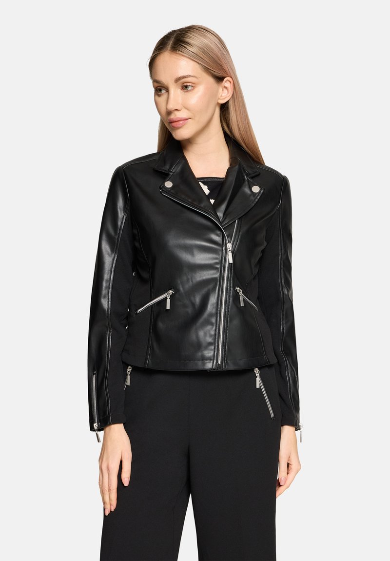 Frau trägt eine taillierte schwarze Lederjacke mit asymmetrischem Reißverschluss und Reißverschlusstaschen, kombiniert mit schwarzer Hose, blickt zur Seite.