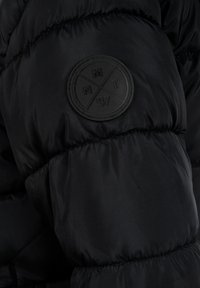 Zwarte pufferjack met een gestructureerde buitenkant en een ronde zwarte rubberen logo-patch op de mouw met cirkelvormige ontwerpen en letters.