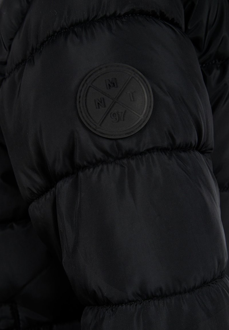 Doudoune noire avec un extérieur texturé et un patch en caoutchouc noir rond sur la manche avec des motifs circulaires et des lettres.