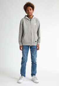Felpa grigia con zip, logo bianco, tasche frontali e cappuccio regolabile con cordino, abbinata a jeans azzurri chiari e sneakers bianche.