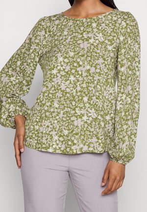 Kvinde iført en grøn og hvid blomstret langærmet bluse med puffede ærmer og lyse lavendelbukser, stående med den ene hånd på hoften.