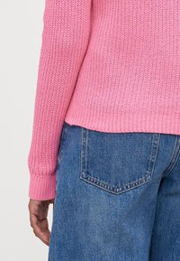 Růžový pletený svetr s žebrovanými manžetami a lemem, kombinovaný s modrými džíny z denimu s zadními kapsami a mírně volným střihem.