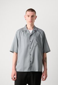 Camisa de botón de manga corta en gris claro, con cuello, un bolsillo en el pecho y textura suave. Lleva puesta una camiseta blanca por debajo.