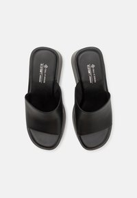 Sandales glissantes véganes noires avec une large sangle supérieure et une semelle intérieure ergonomique. Texture lisse, semelle plate et design minimaliste.