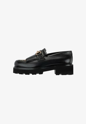 Zwarte leren loafer met dikke zool, gouden gesp-detail en franjes op een witte achtergrond.