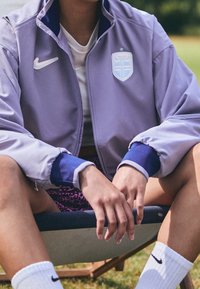 Nike purple zip-up jacket with a blue and white logo, featuring contrasting cuffs and a white inner collar, paired with patterned shorts.