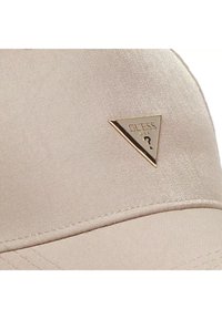 Beżowa czapka o gładkiej teksturze materiału, z złotym trójkątnym logo z napisem "GUESS USA." Akcentowane szwy wzdłuż daszka.