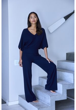 Marineblauer Strick-Jumpsuit mit V-Ausschnitt, weiten Ärmeln und locker geschnittenen, weiten Hosenbeinen, ergänzt durch eine taillierte Passform. Das Model steht auf Betontreppen.