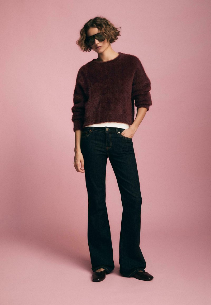 Burgunderfarbener, flauschiger Strickpullover im cropped Stil, kombiniert mit dunkelblauen Schlagjeans. Das Model trägt schwarze oversized Sonnenbrillen und braune Ballerinas.