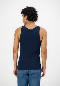 Tricou fără mâneci, de culoare albastru marin, cu decolteu curbat și ajustat, fabricat dintr-un material moale. Purtați cu blugi de culoare albastru deschis, văzut din spate.